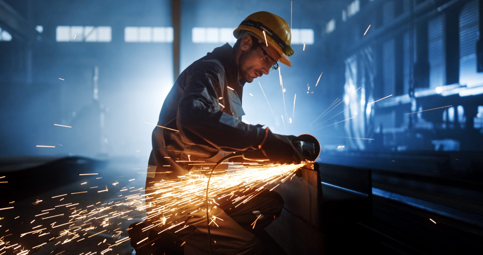 A worker in protective gear uses a grinder on a metal beam, producing bright sparks in an industrial factory setting.