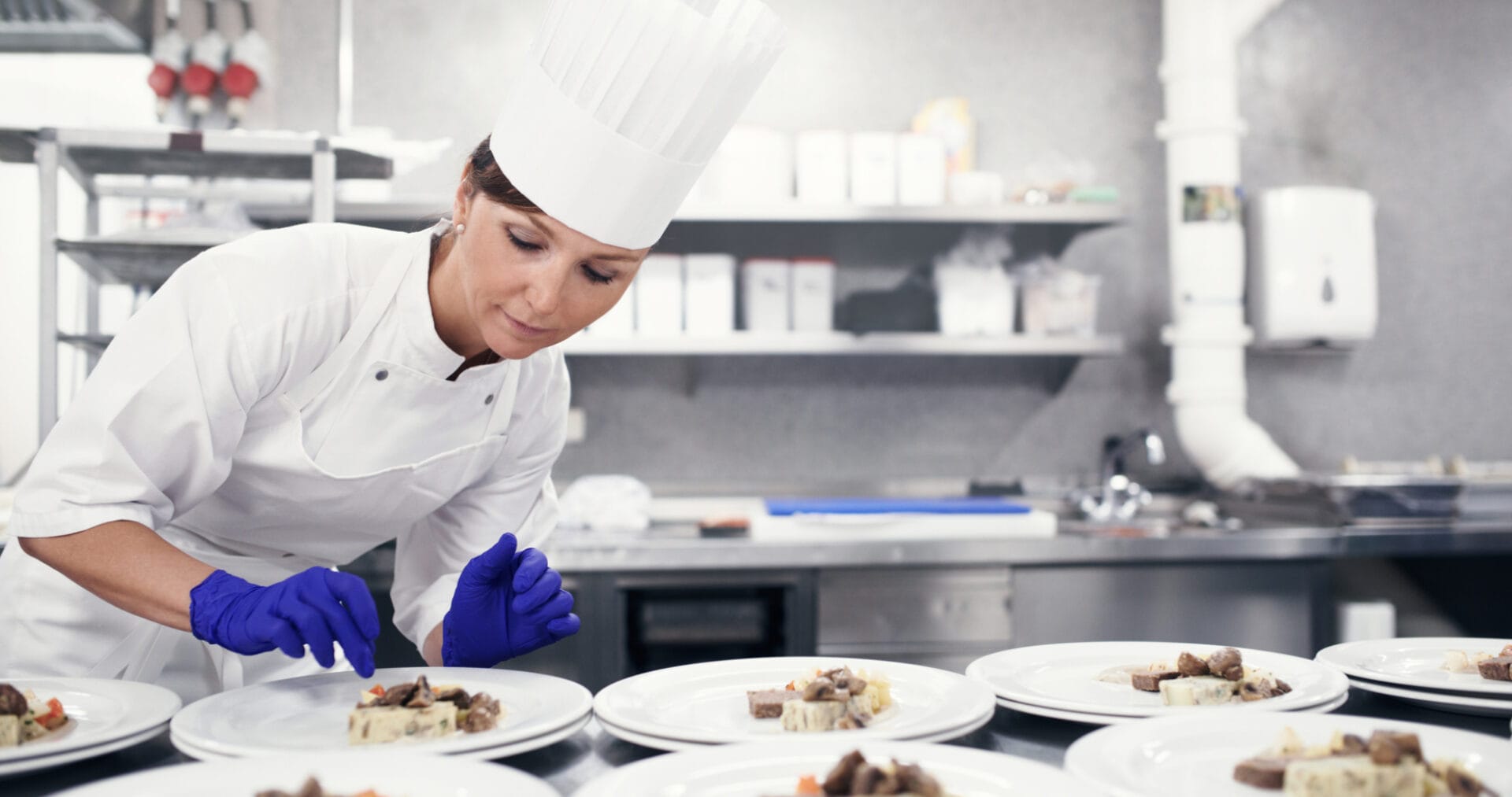 A chef in a white uniform and hat arranges food on plates in a commercial kitchen, wearing blue gloves.